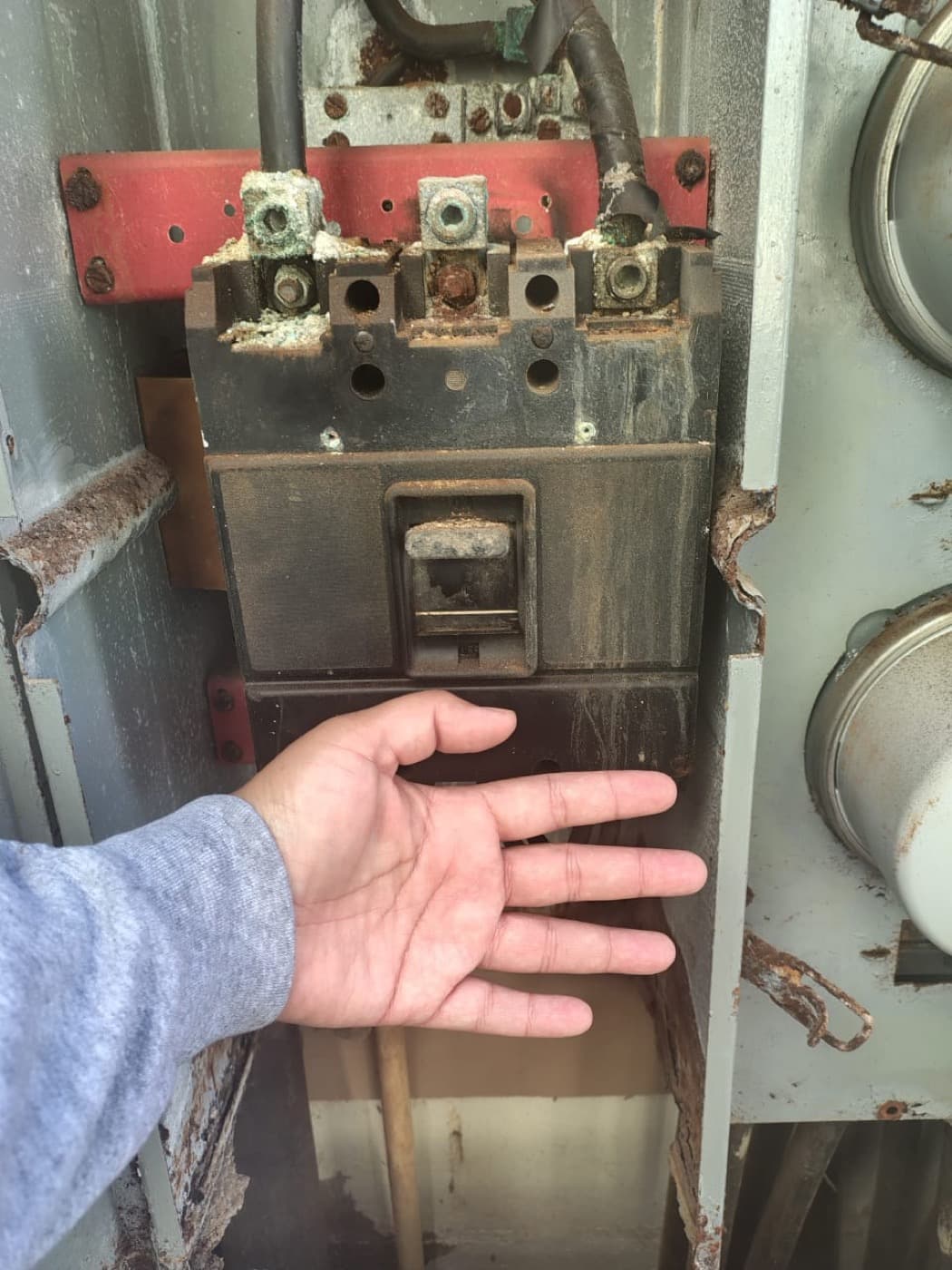 Close-up of a heavily corroded main breaker with a hand for scale, showing rust, copper corrosion, and burn marks